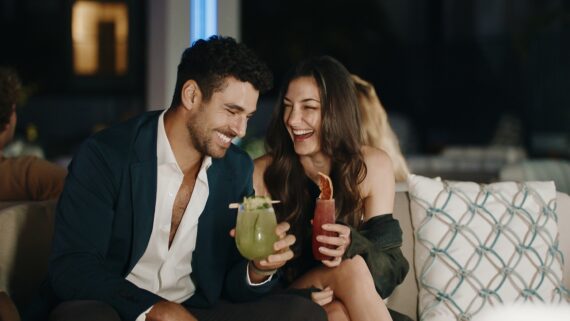 A man and woman sit close together on a couch, smiling and laughing while holding colorful cocktails, enjoying a night out in a stylish, dimly lit setting.