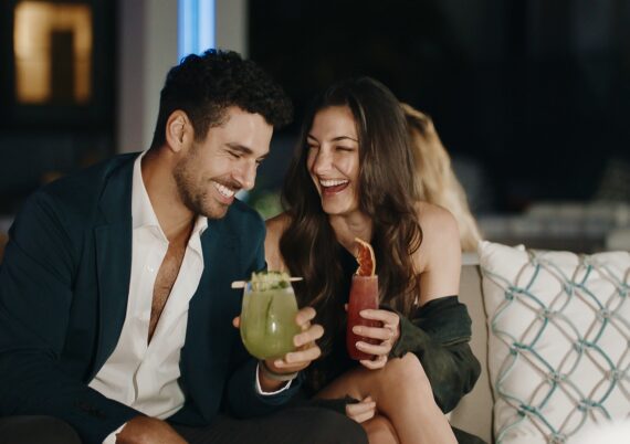 A man and woman sit close together on a couch, smiling and laughing while holding colorful cocktails, enjoying a night out in a stylish, dimly lit setting.