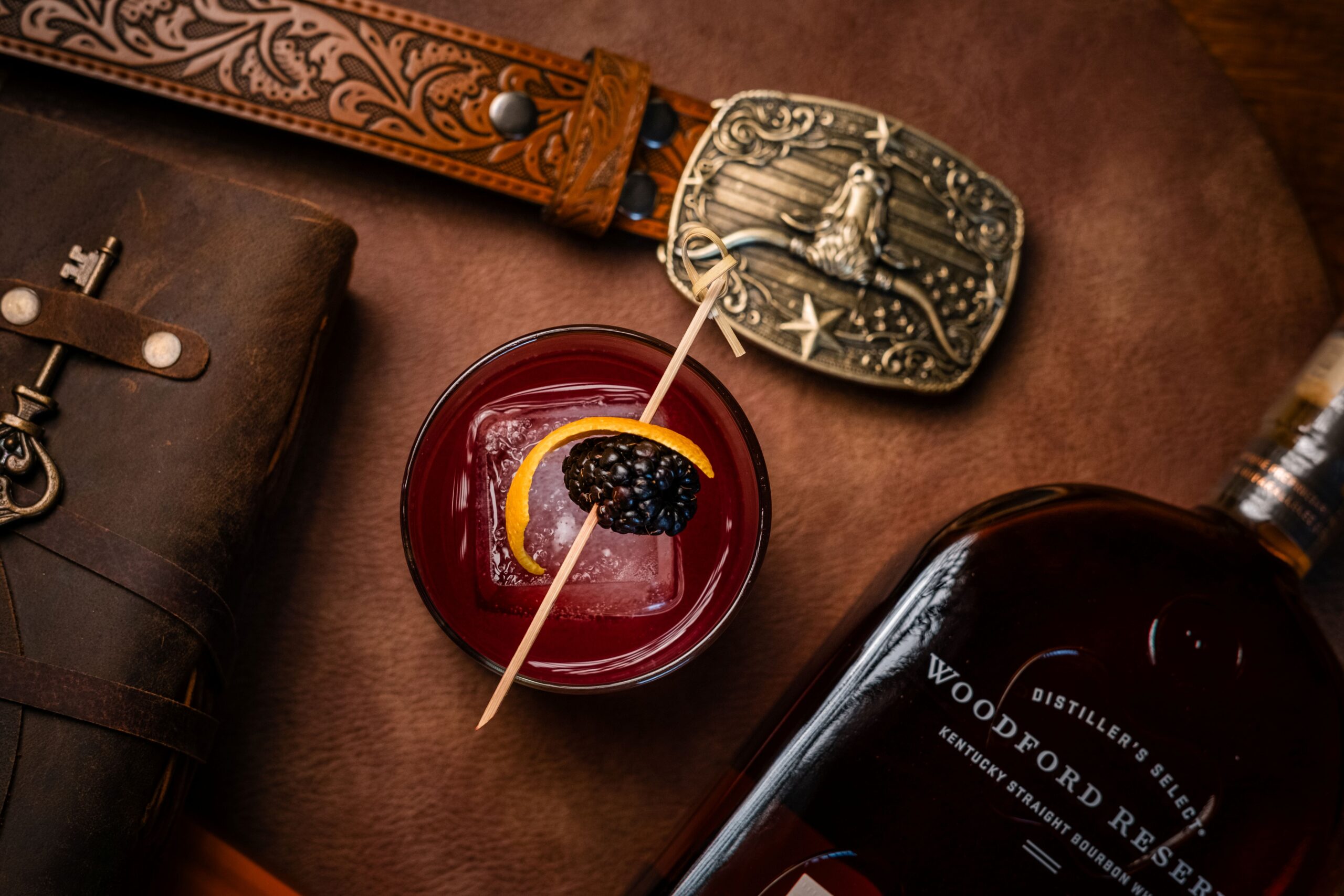 A cocktail with ice, lemon twist, and blackberry garnish is on a brown surface beside a leather belt with a decorative buckle, a leather-bound book, and a bottle labeled Woodford Reserve.