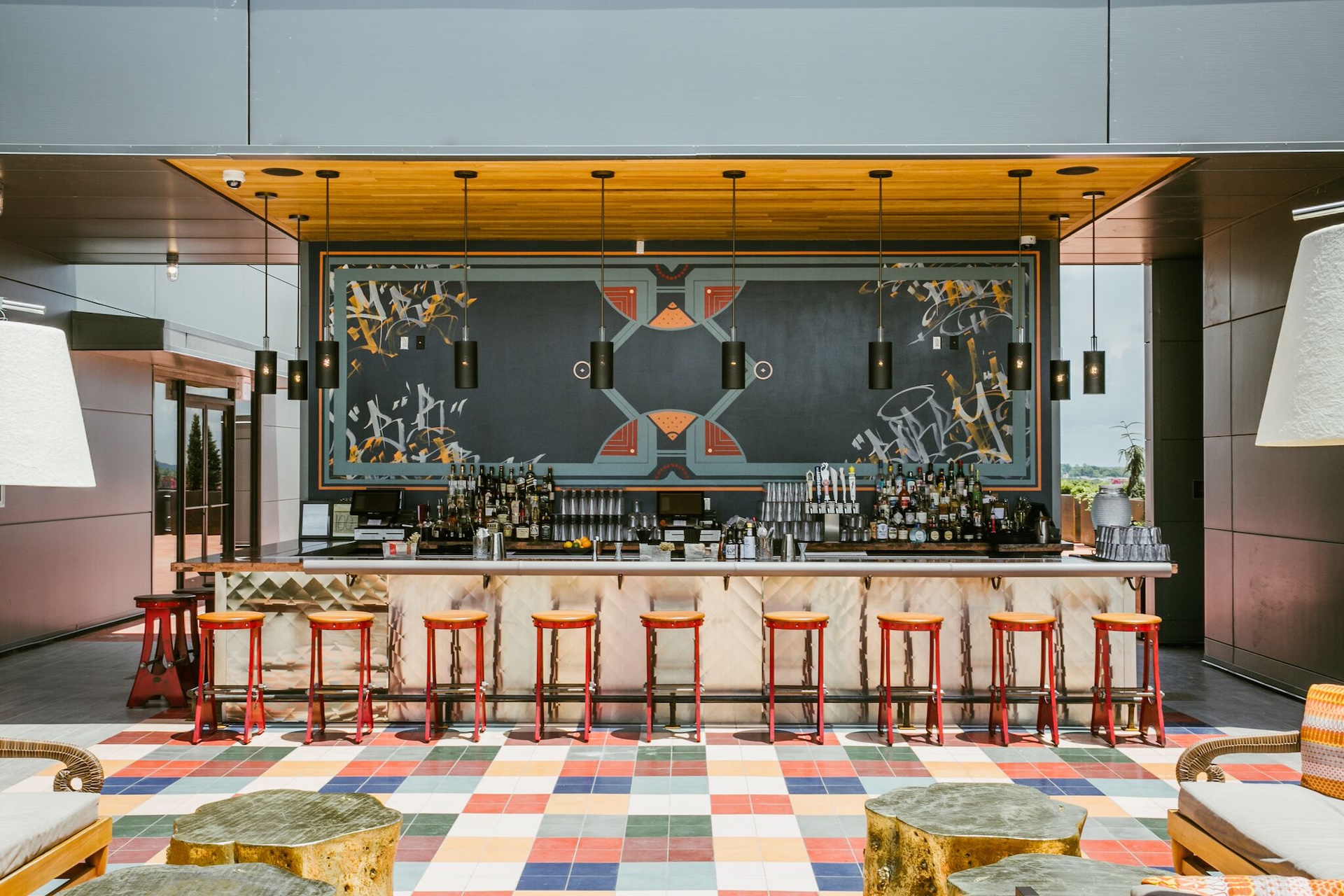 A modern bar with a marble counter, red stools, a large geometric mural, hanging pendant lights, and colorful tiled flooring. Some wooden log tables and chairs are in the foreground.