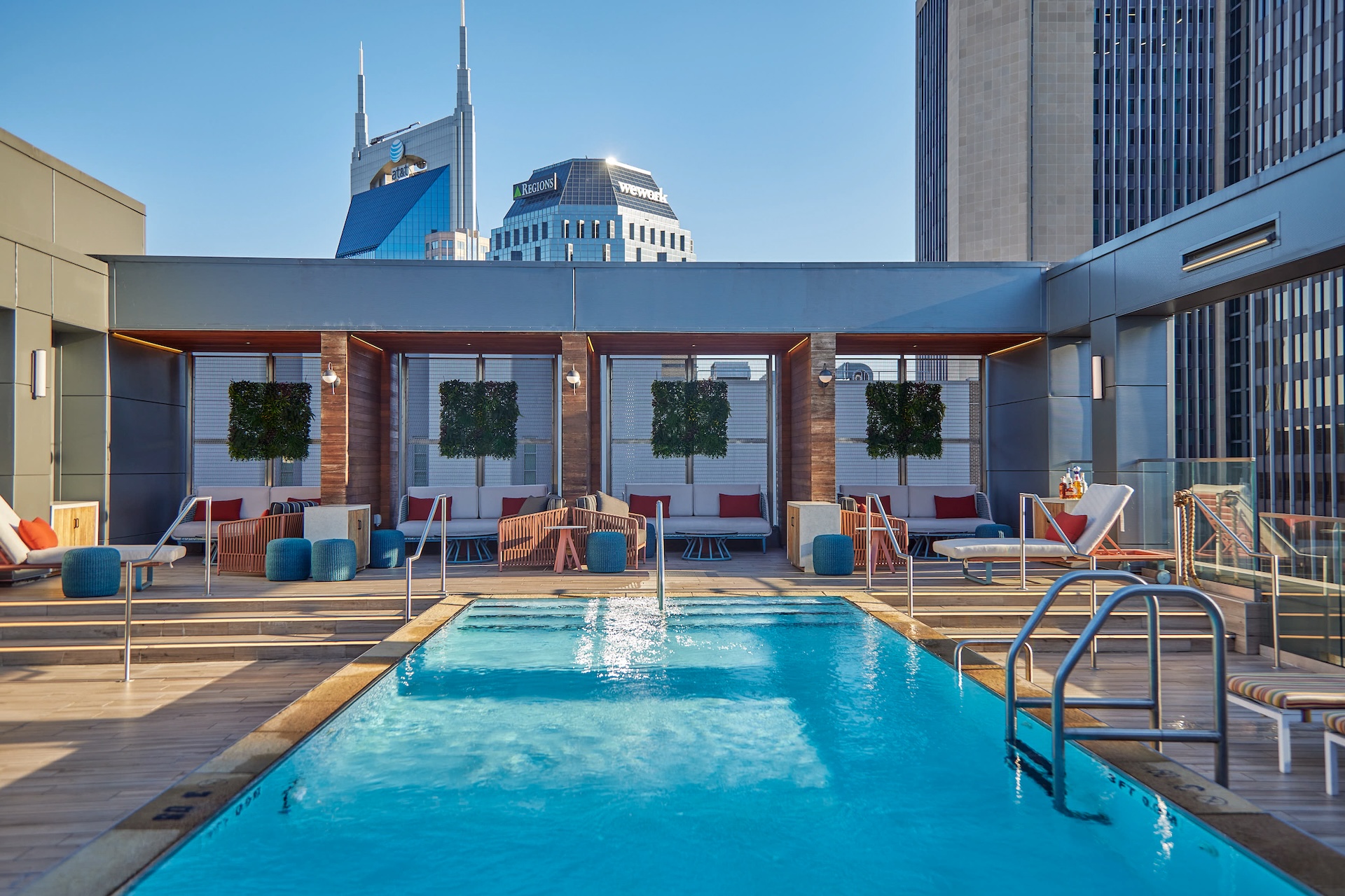 Rooftop pool with modern lounge seating, wooden cabanas, and city skyscrapers in the background under a clear blue sky.