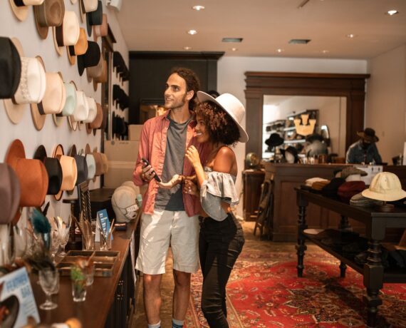 A couple stands in a hat shop, smiling and trying on hats. The man holds a light-colored hat, while the woman hugs him from the side. The shop is filled with hats displayed on the wall and tables.