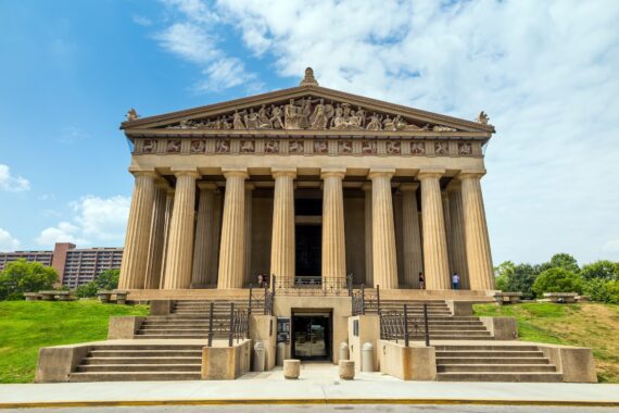 The image shows the Parthenon replica in Nashville, Tennessee, featuring tall columns, detailed pediment sculptures, and stone steps, with a partly cloudy sky and greenery in the background.