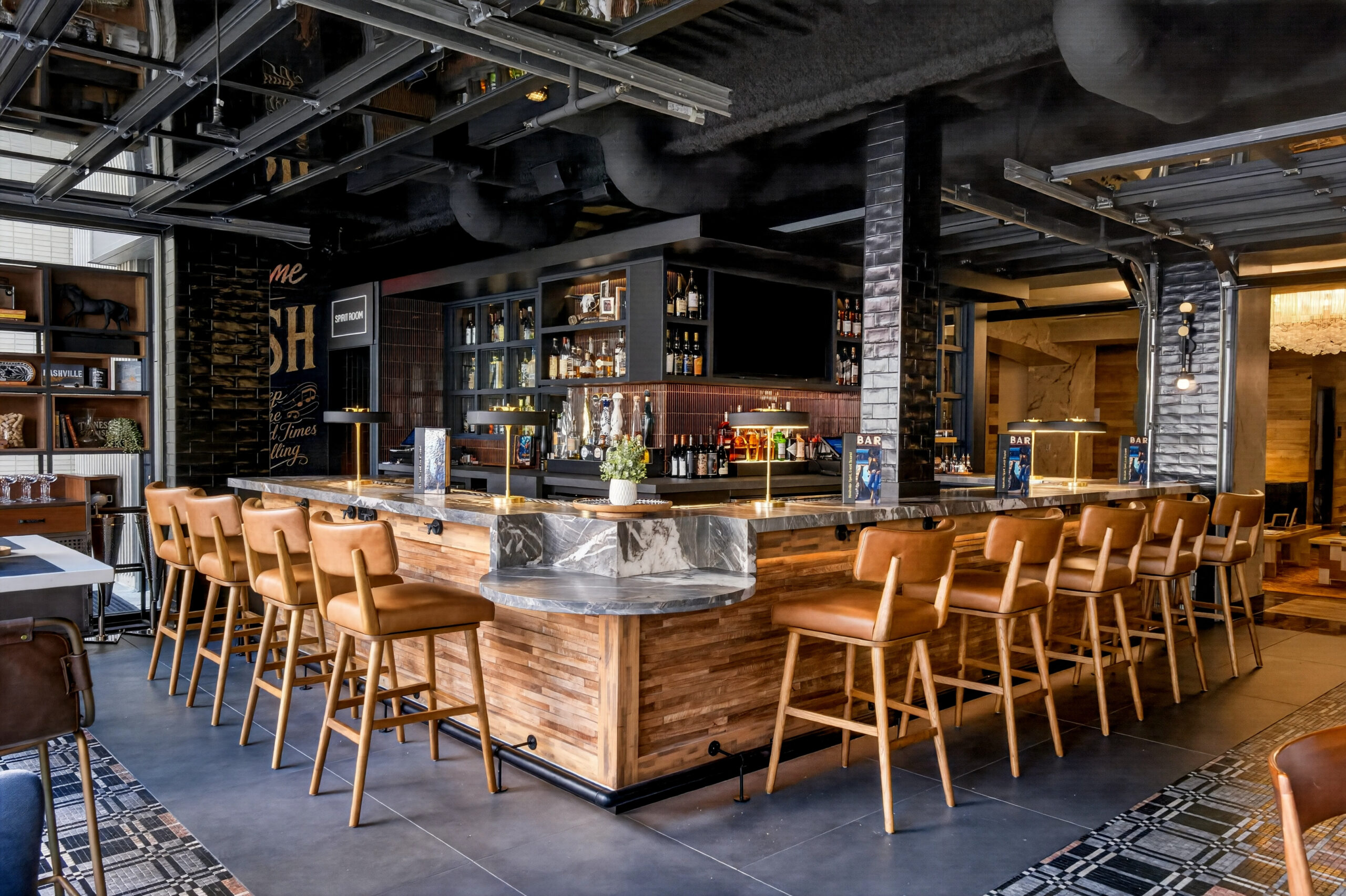 A modern bar with wooden stools, a marble countertop, shelves of liquor bottles, and industrial decor. The space is well-lit, with tiled floors and a mix of wood and black metal accents.