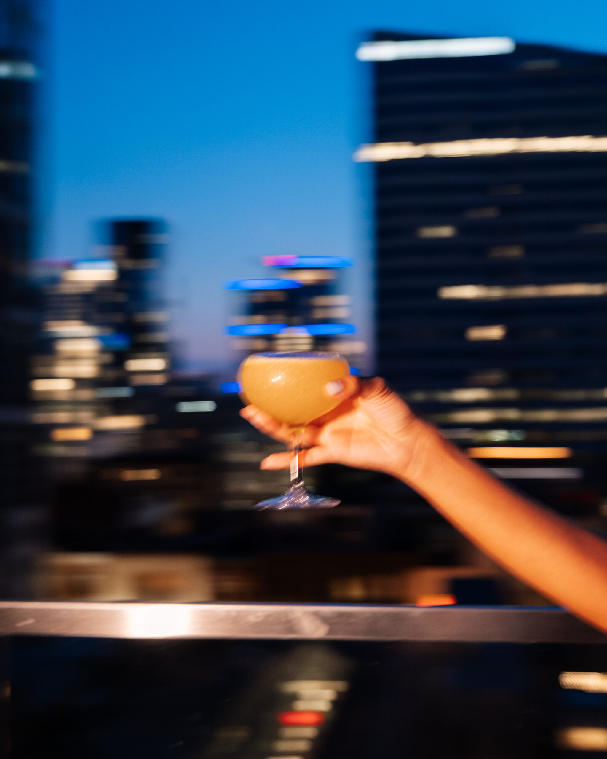A hand holds a cocktail glass against a blurred city skyline at twilight, with buildings lit up in the background.