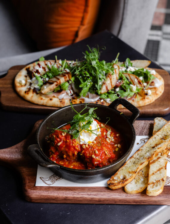 A skillet of meatballs in tomato sauce with cheese and herbs, served with toasted bread slices, sits in front of a flatbread topped with greens and grilled chicken on a wooden board.