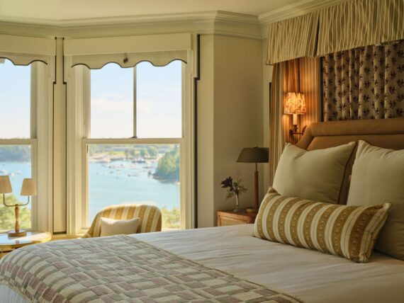 A cozy summer house-inspired bed in one of the guest rooms at The Asticou with a view of the harbor in the background.