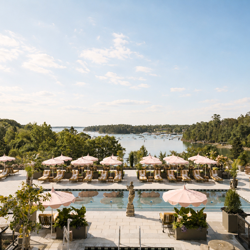 A luxurious outdoor pool area with pink umbrellas and lounge chairs overlooks a calm river surrounded by lush greenery and boats under a clear, sunny sky.