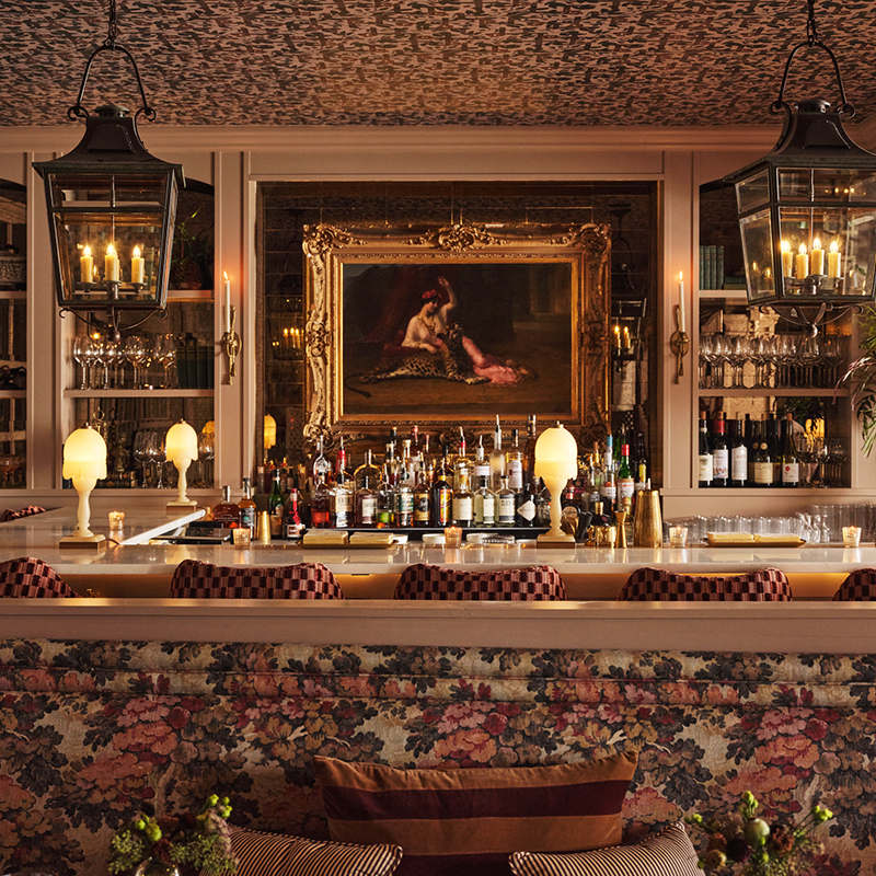 A warmly lit bar with floral-patterned decor, vintage lamps, hanging lanterns, and a gold-framed painting above shelves of bottles and glassware. The chairs have a checkered pattern and the atmosphere feels cozy and elegant.