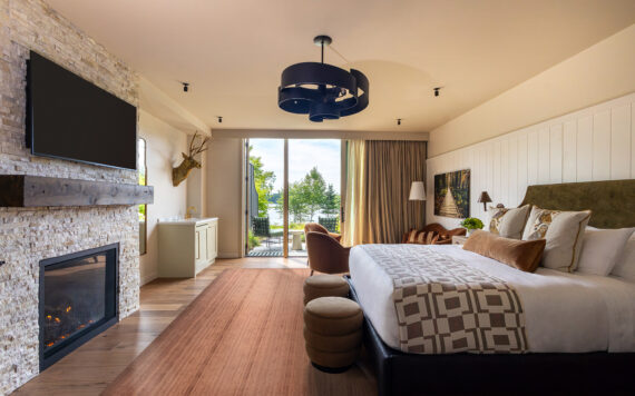 Modern hotel room with a king bed, fireplace, wall-mounted TV, large window doors opening to a balcony with trees and water view, contemporary lighting, and stylish decor.