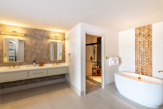 Modern bathroom with double sinks, wall-mounted mirrors, gold fixtures, a freestanding white bathtub beside a textured accent wall, soft lighting, and an open door leading to a walk-in shower area.
