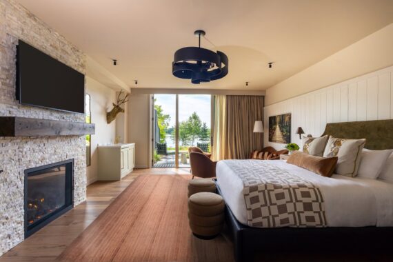 Modern hotel room with a king bed, fireplace, wall-mounted TV, seating area, large window with a lake view, and neutral-toned decor.