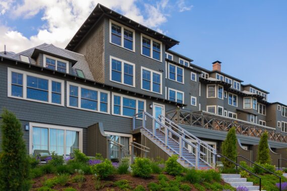 A large, modern gray building with many windows and balconies, set against a blue sky. There are stairs, green shrubs, and purple flowers in the landscaped front yard.