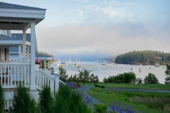 White porch overlooking a scenic bay with sailboats, green grass, and colorful flowers in the foreground. Mist rises over a forested hill across the water under a blue sky.