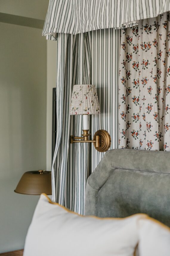 A brass wall lamp with a floral-patterned shade is mounted beside a bed with a gray upholstered headboard and white pillow. Striped and floral curtains frame the background.