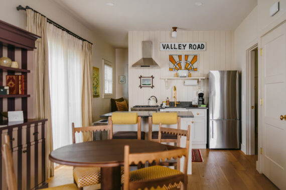 A cozy kitchen and dining area with wooden furniture, a round table, yellow cushioned chairs, stainless steel fridge, and a Valley Road sign above the sink. Sunlight streams in through sheer curtains.