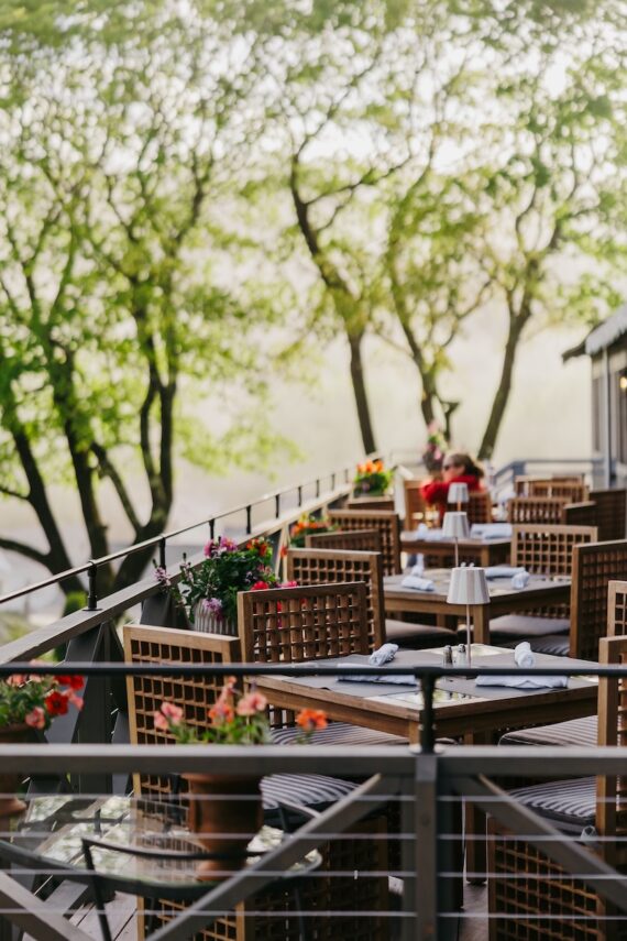 Outdoor restaurant patio with wooden tables and chairs, set for dining, surrounded by potted flowers and lush green trees, creating a serene and inviting atmosphere.
