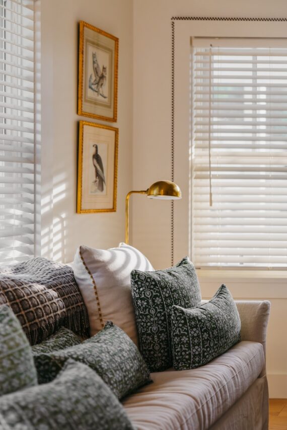 Sunlit living room with a beige couch, green patterned pillows, framed bird art on the wall, a gold floor lamp, and white blinds casting striped shadows.