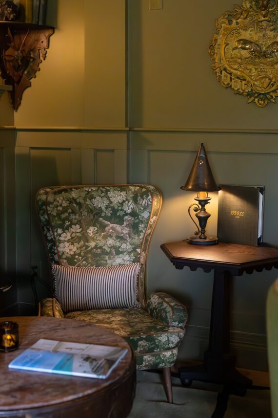 A cozy green floral armchair with a striped pillow sits by a small wooden table holding a lamp and book, in a warmly lit room with green paneled walls and ornate wall decor.