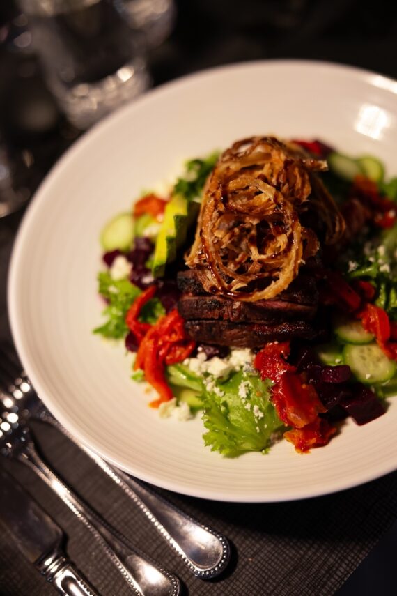 A plate of salad topped with grilled steak strips, crispy fried onions, lettuce, cucumbers, red peppers, beets, and crumbled cheese, set on a table with a fork, knife, and spoon.