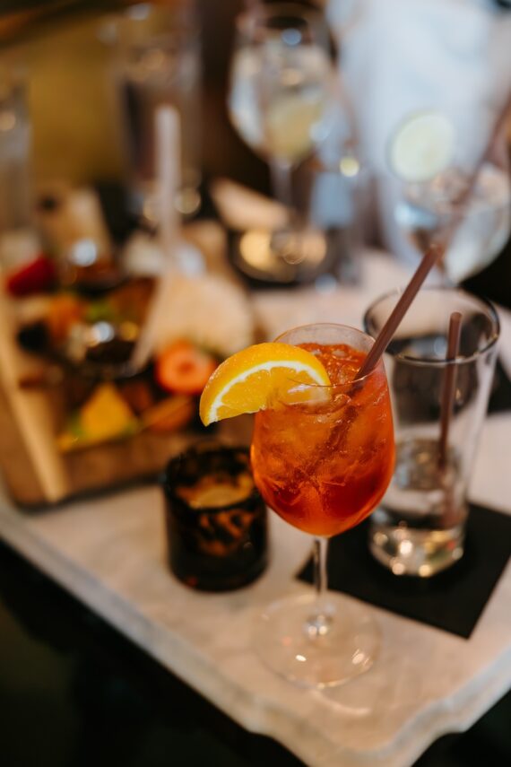 A glass of orange cocktail with ice, garnished with orange slices and a straw, sits on a table next to an empty glass, a candle, and a wooden platter of assorted appetizers.