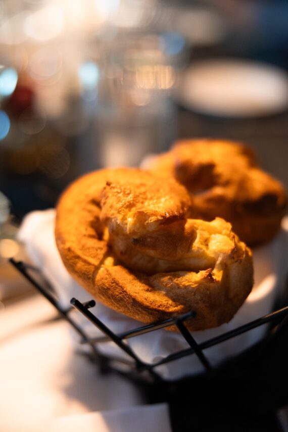Two golden-brown popovers rest in a black wire basket lined with a white cloth, with a softly blurred background of glassware and plates.