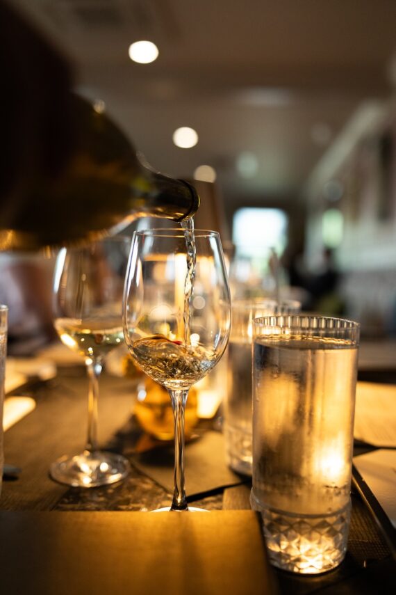 A close-up of white wine being poured into a glass on a dimly lit restaurant table, surrounded by water glasses and menus.