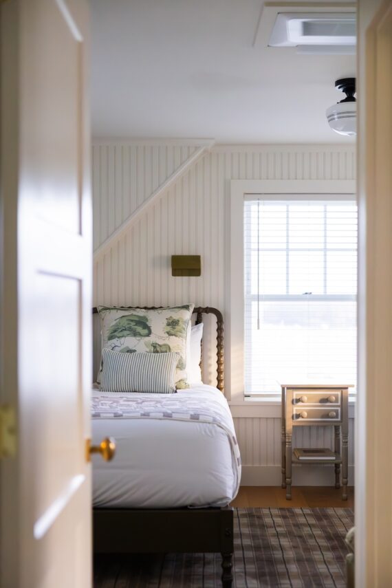A bright bedroom with a single bed, floral and striped pillows, a small nightstand, and a window with white blinds letting in natural light.