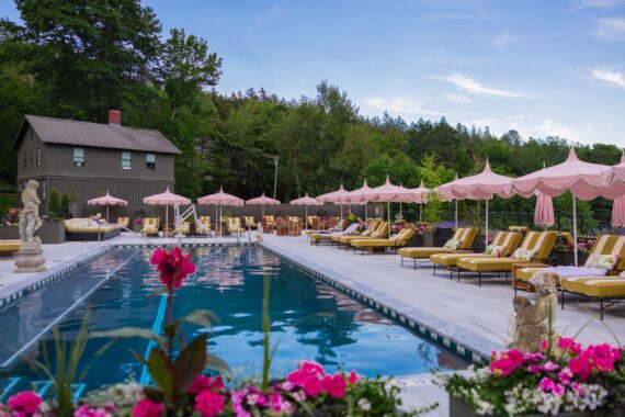 A luxurious outdoor pool with striped lounge chairs, pink umbrellas, and statues, surrounded by flowers and greenery. A rustic house is in the background under a blue sky.