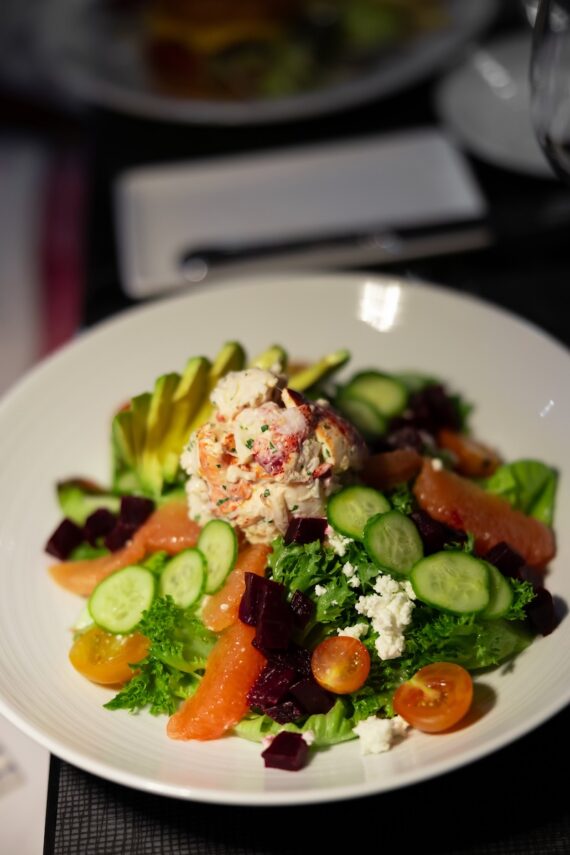 A colorful salad with lettuce, sliced cucumber, cherry tomatoes, avocado, beets, feta cheese, grapefruit segments, and a scoop of creamy seafood salad in the center, served on a white plate.