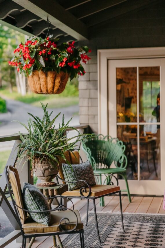 A cozy porch with cushioned chairs, a small table, potted plants, a hanging basket of red flowers, and glass doors leading inside.