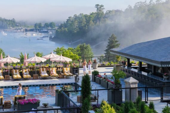 A poolside scene at a resort overlooking a misty bay with boats. People relax on lounge chairs and walk by the pool, surrounded by flowers and greenery, with forested hills in the background.