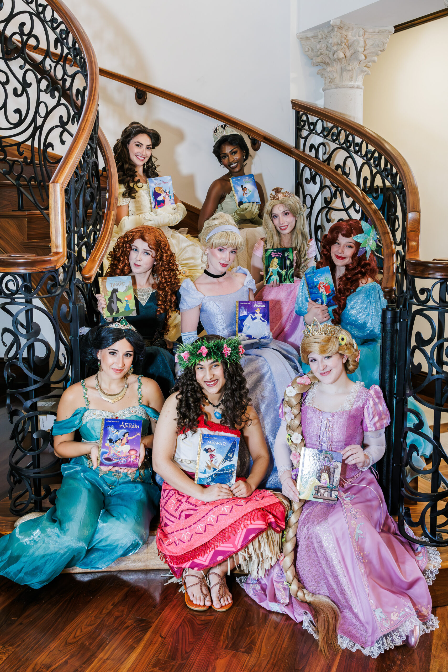 Nine women dressed as various fairytale princesses sit on a staircase with ornate black railings, each holding a matching storybook. The setting is indoors, with a polished wooden floor and white columns in the background.