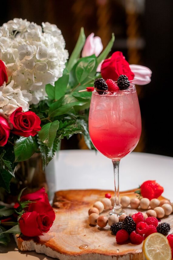 A pink cocktail with ice and berries is served in a wine glass, placed on a wooden board with assorted berries, lemon slices, and white beads. A bouquet of red and white flowers is in the background.