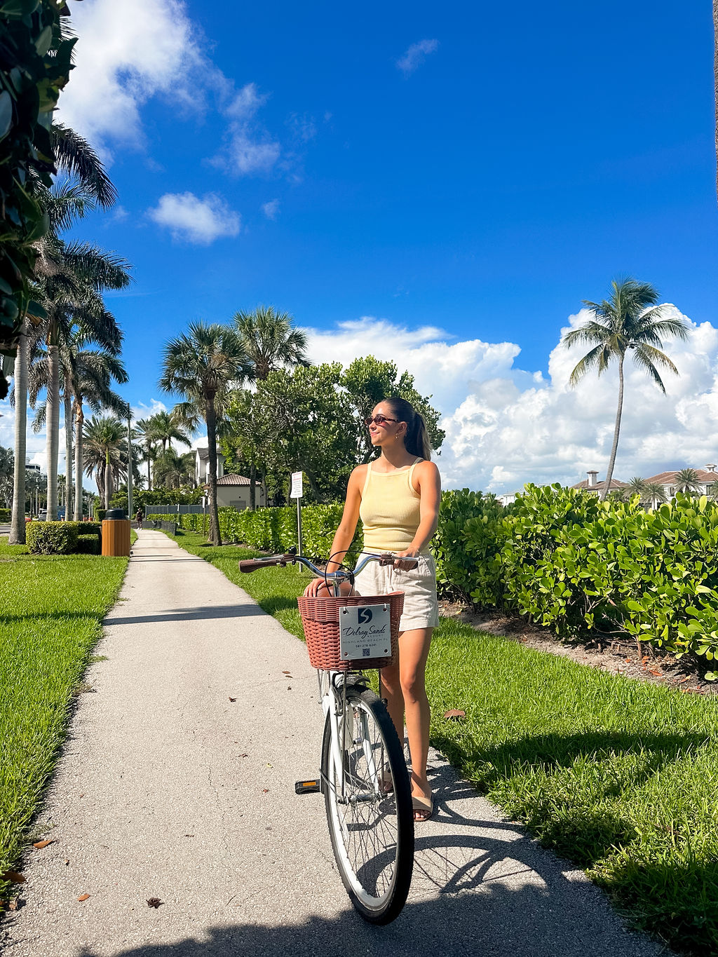 A woman wearing sunglasses and a yellow top walks a bicycle along a sunny, palm tree-lined path with bright green grass under a clear blue sky.