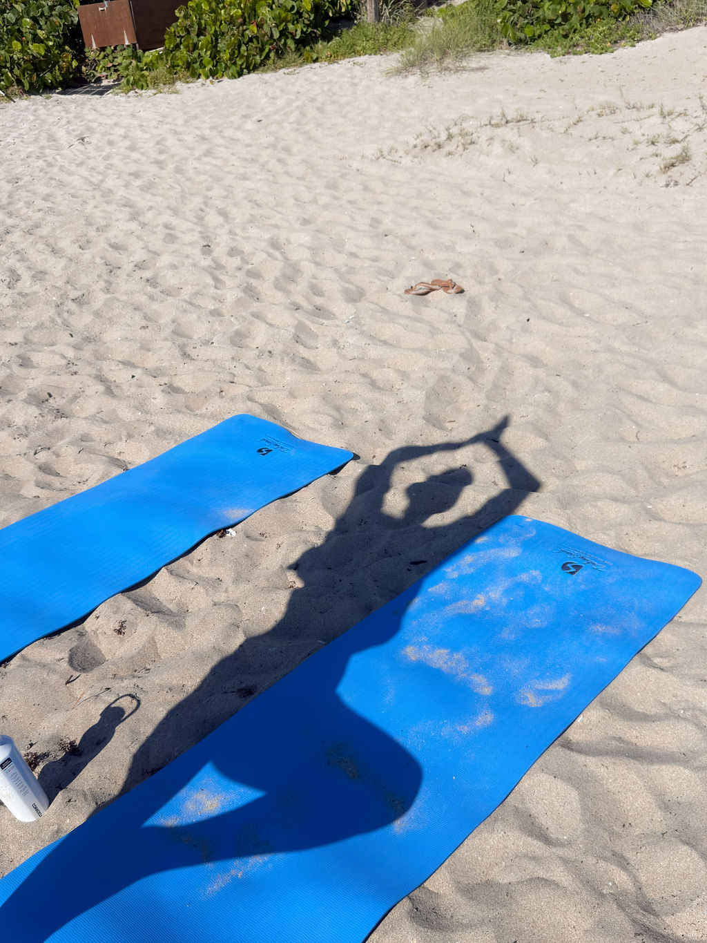 A shadow of a person doing a yoga pose appears on a blue mat on sandy beach, next to another empty mat. Some greenery is visible in the background.
