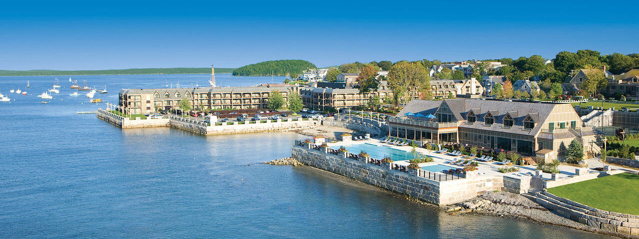 Aerial view of a coastal resort with buildings and a swimming pool by the water. Sailboats are on the ocean and lush green trees surround the area under a clear blue sky.