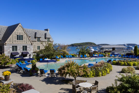A scenic view of a hotel with a stone facade, surrounded by a pool area with blue lounge chairs and umbrellas. In the background, there is a clear blue sky and a glimpse of the ocean with boats and distant islands.