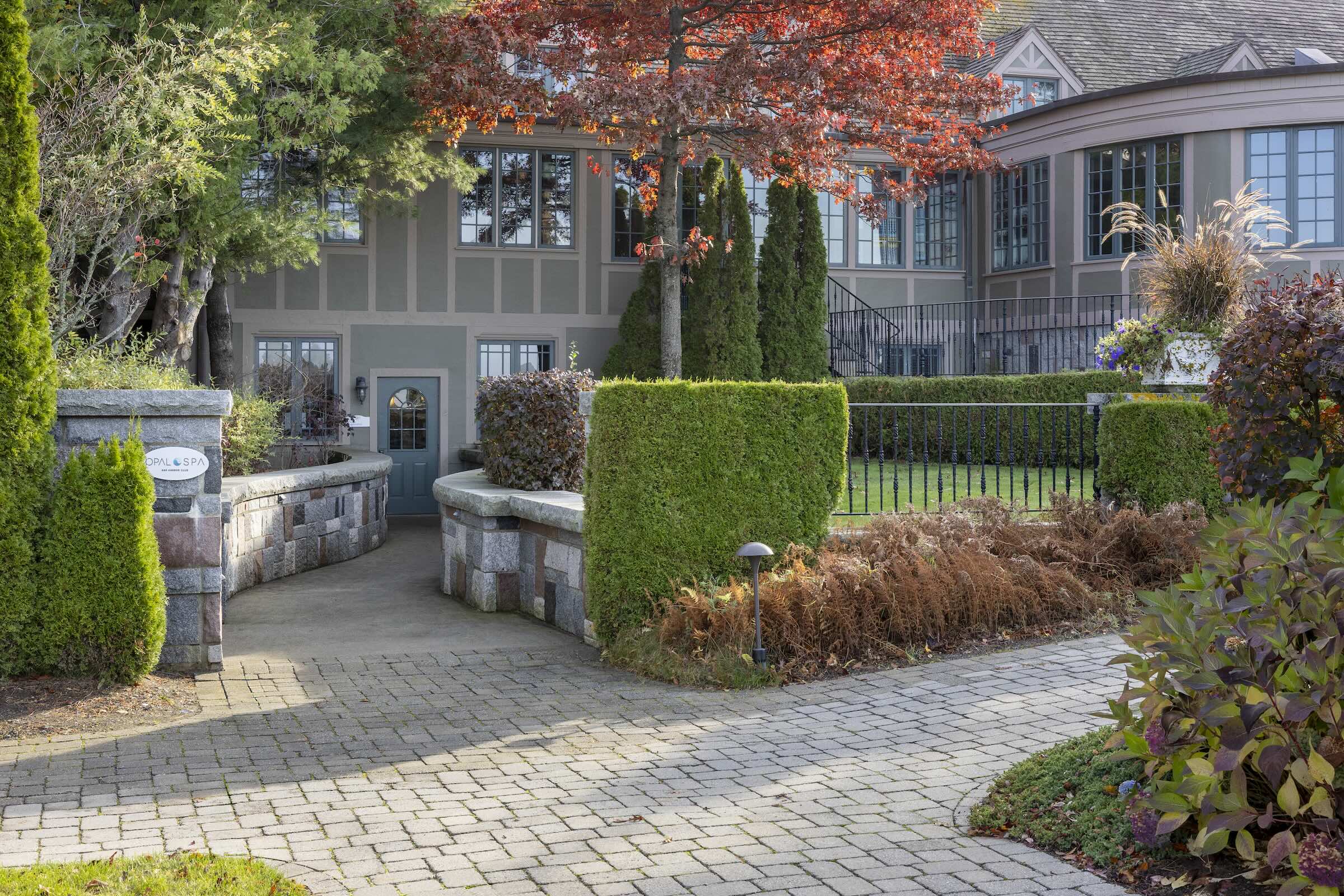 Pathway with stone paving leading to a large house surrounded by manicured hedges and colorful autumn trees. The house has multiple windows and a slightly curved entrance, with a mix of brick and siding on its facade.
