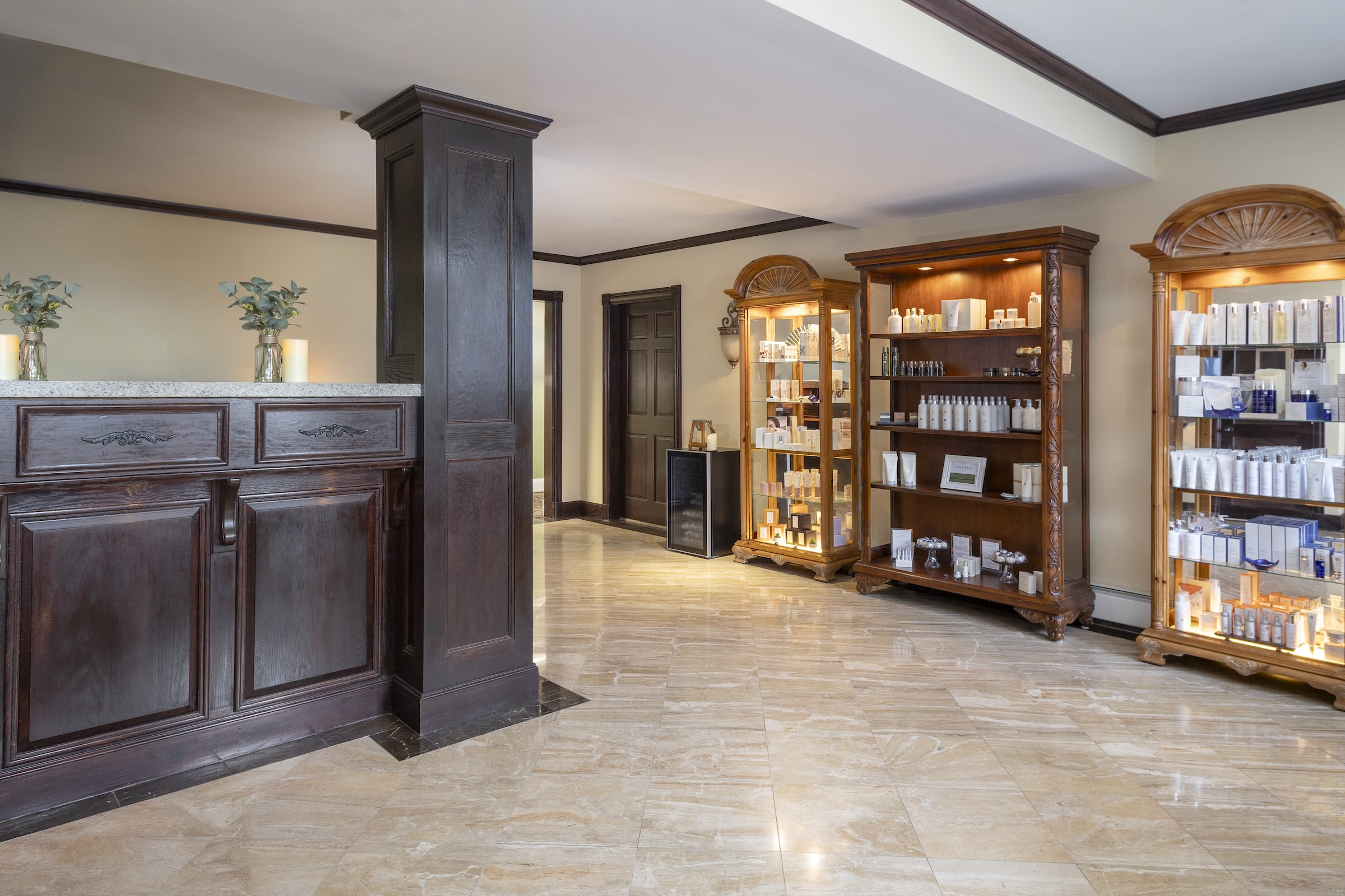 A bright, elegant spa reception area with polished marble floors, a dark wooden reception counter on the left, and two illuminated shelving units displaying skincare products on the right. Two potted plants adorn the counter.