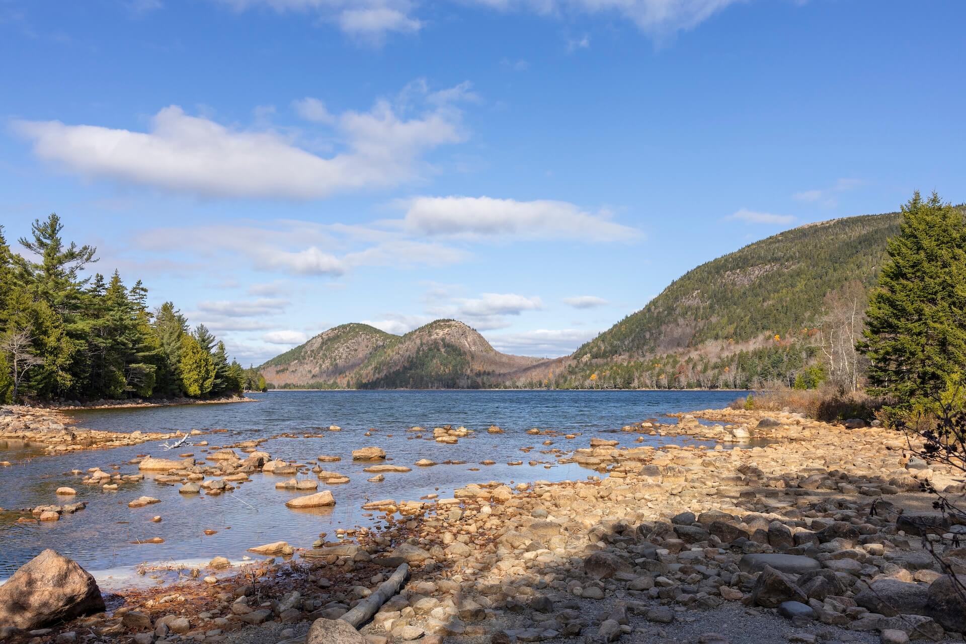 A clear, shallow lake with scattered rocks along the shoreline, surrounded by forested hills and mountains under a blue sky with scattered clouds.