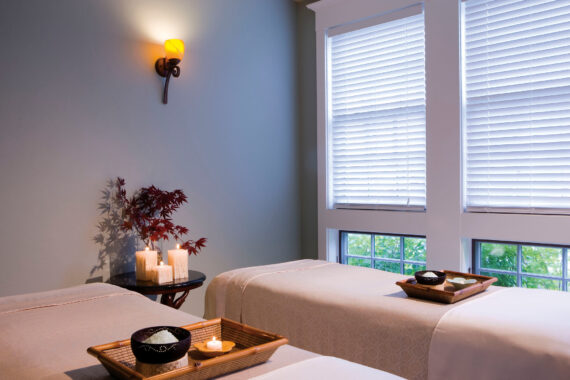 A peaceful spa room with two massage tables, each topped with a wooden tray holding a bowl and candle. Soft natural light filters in through white blinds; candles and a plant decorate a corner table.