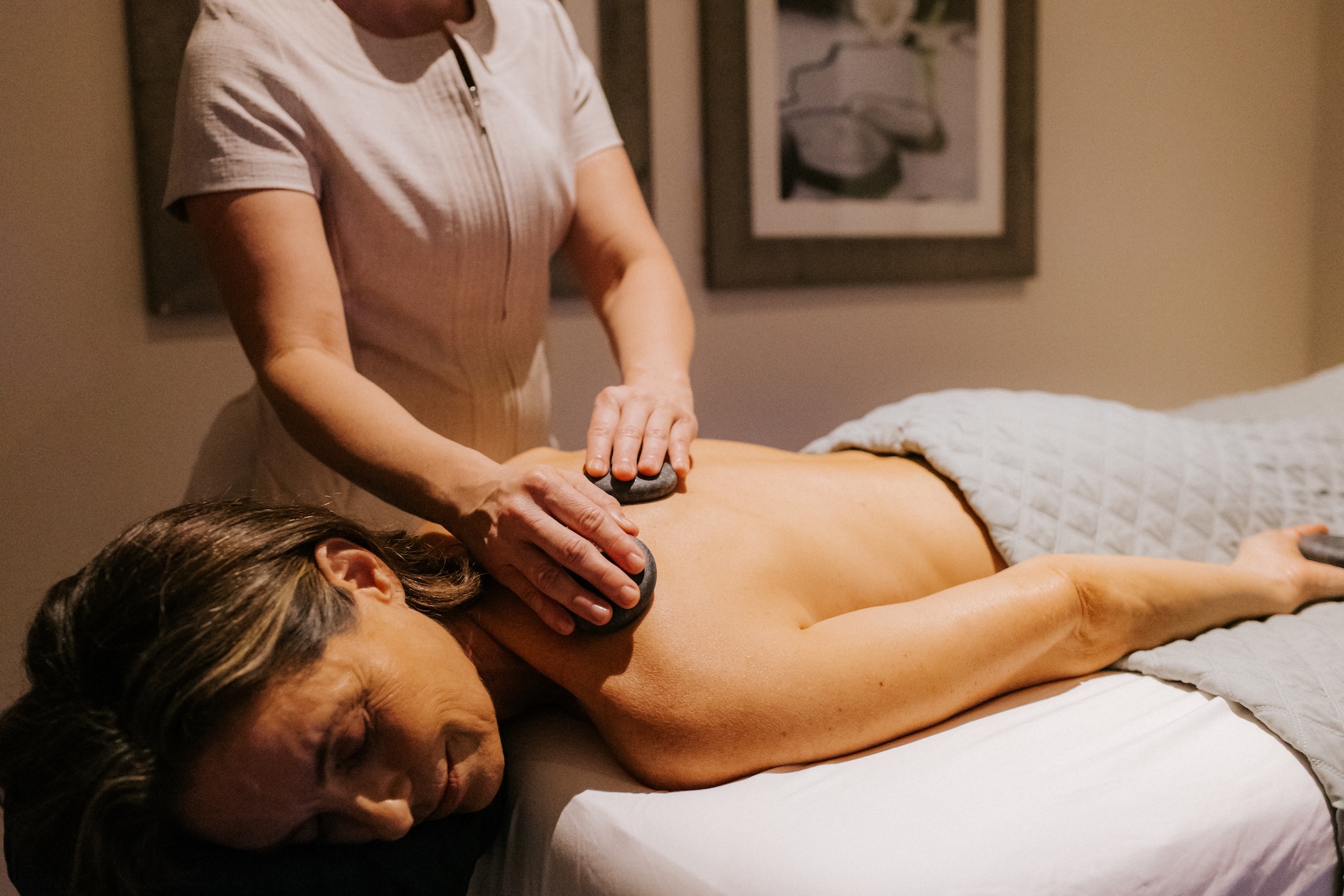A person receiving a hot stone massage on their back, lying face down on a massage table. Theyre covered with a gray quilt. The massage therapists hands are visible placing stones on the persons back in a softly lit room.