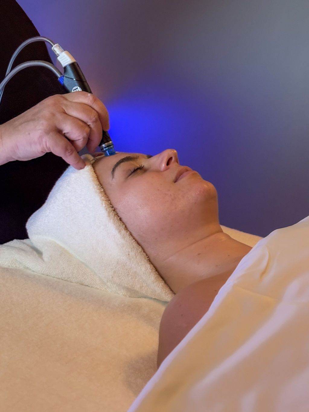 A person lies on a spa bed with eyes closed, covered in a towel and blanket, while a hand uses a skincare device on their forehead under soft blue lighting.