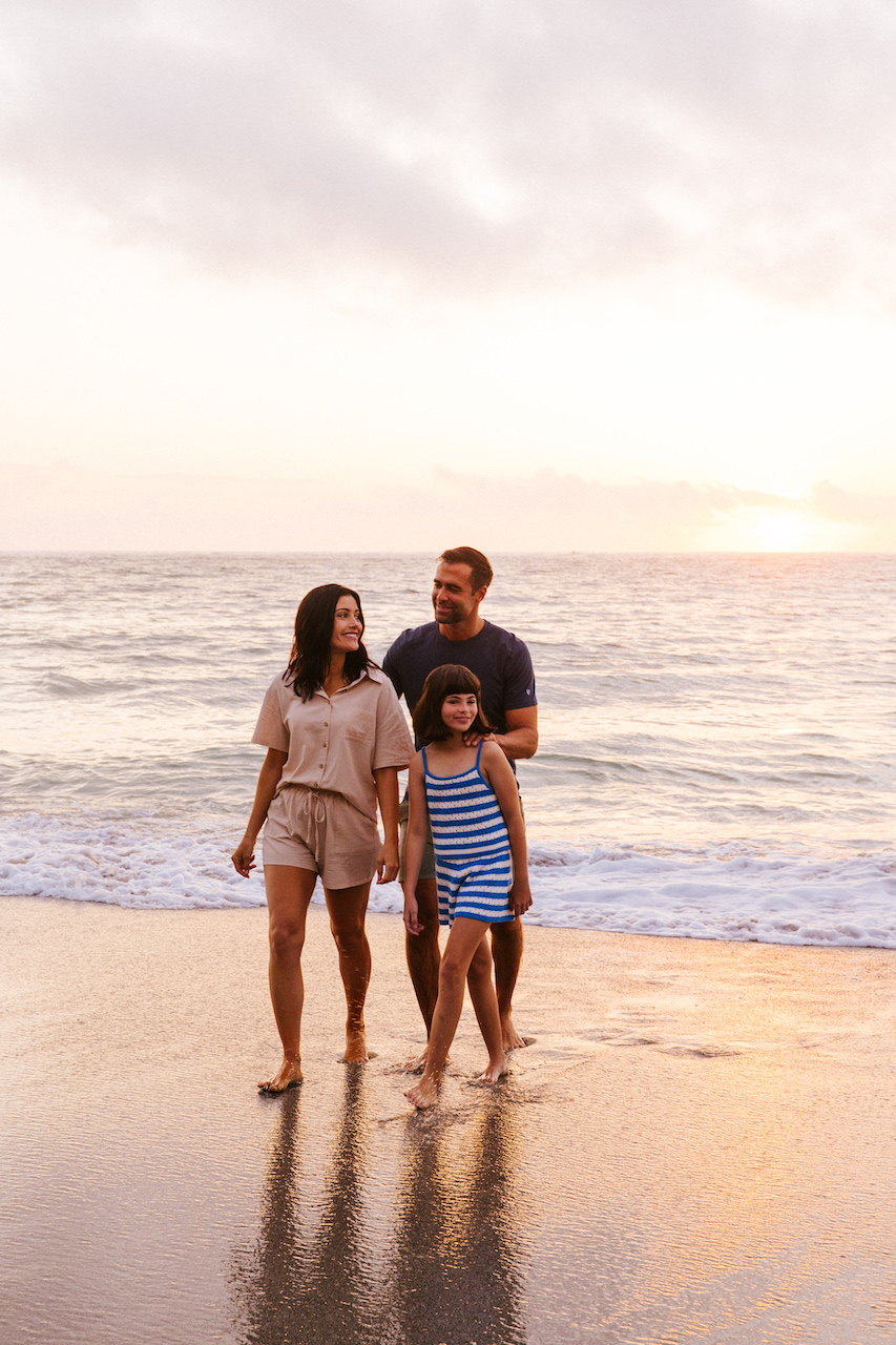 A family of three walks barefoot along the shoreline at sunset. A woman in beige attire, a man in a navy shirt, and a girl in a blue striped dress hold hands, enjoying the serene beach as the sun sets over the ocean.