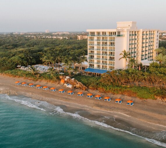 Aerial view of a beachfront hotel with a modern building. Orange and blue umbrellas line the sandy beach next to gentle ocean waves. Lush greenery surrounds the area, and a hazy horizon is visible in the background.