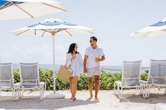 A couple walks on a sandy beach, each holding a drink. They are under beach umbrellas, with empty loungers nearby. The sky is clear, and the ocean is in the background. Both are casually dressed, enjoying a sunny day.