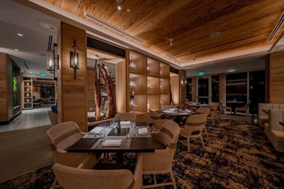 A modern restaurant interior with wooden accents, soft lighting, woven chairs, set tables, and a decorative wall partition. The atmosphere is warm and inviting, with a mix of neutral and earthy tones.