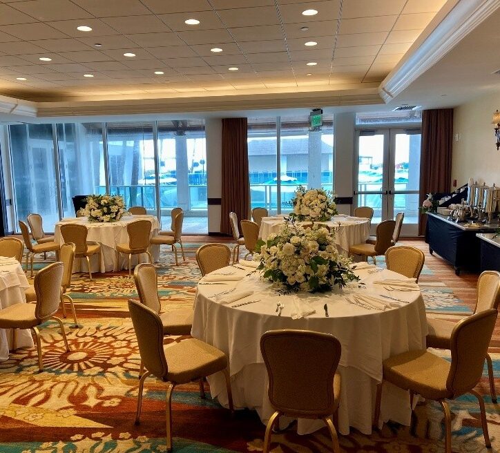 Elegant banquet room with round tables covered in white tablecloths, each decorated with large white floral centerpieces. Beige chairs surround the tables, and large windows provide natural light and views outside.