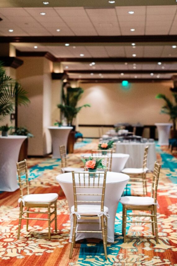 A banquet hall with round tables covered in white cloths, gold chairs, floral centerpieces, and cocktail tables. The room has colorful carpet, potted plants, and soft ceiling lights, creating an elegant event setting.